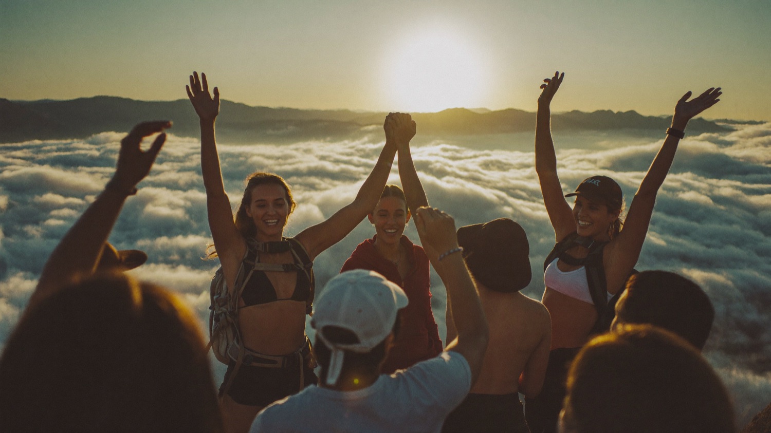 A group reaching a mountain summit together at sunrise, arms raised